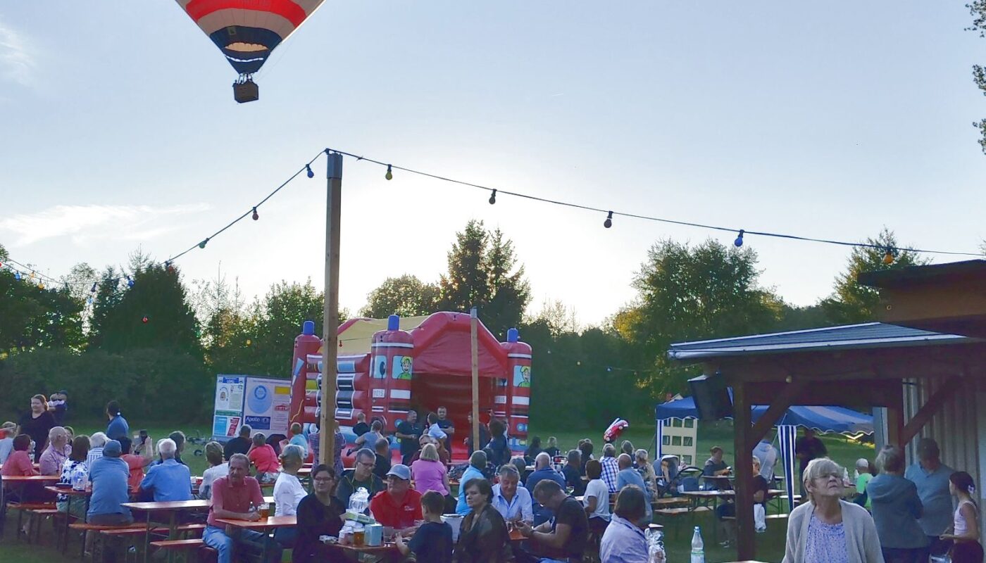 Auf einer Wiese stehen viele Biertischbänke. Zahlreiche Menschen sitzen darauf und feiern. Darüber fliegt ganz tief ein Heißluftballon.