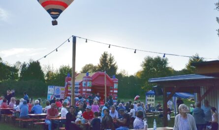 Auf einer Wiese stehen viele Biertischbänke. Zahlreiche Menschen sitzen darauf und feiern. Darüber fliegt ganz tief ein Heißluftballon.