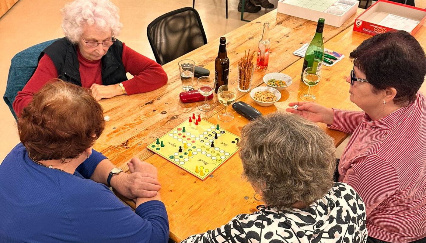 Vier Personen sitzen an einem Tisch. Sie spielen Mensch-Ärgere-Dich-Nicht. auf dem Tisch liegt eine Spielplatte für das Spiel. Daneben stehen verschiedene Getränke und Knabbereien.