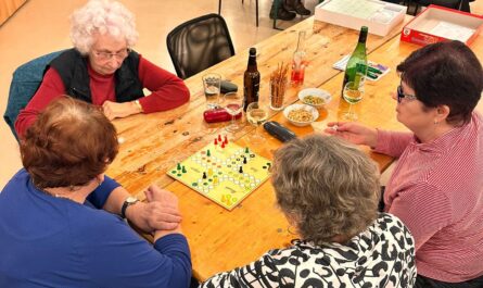 Vier Personen sitzen an einem Tisch. Sie spielen Mensch-Ärgere-Dich-Nicht. auf dem Tisch liegt eine Spielplatte für das Spiel. Daneben stehen verschiedene Getränke und Knabbereien.